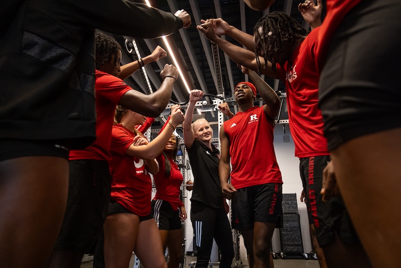 image shows a woman strength coach huddling with a team in the weight room