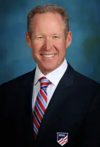 image shows a headshot of a man wearing a black blazer and red and blue striped tie
