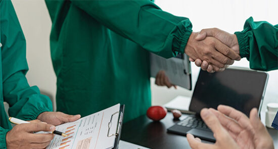 image shows two people in green jackets shaking hands and someone else holding a clipboard with documents