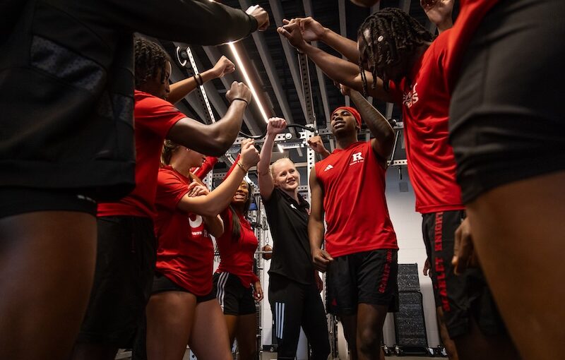 image shows a woman strength coach huddling with a team in the weight room