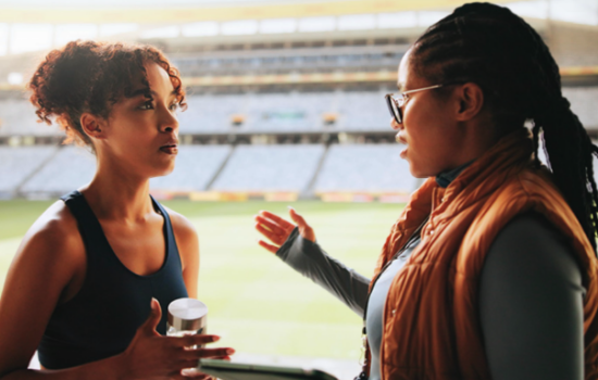 image shows two Black women, one athlete and one coach, talking in the bleachers of a stadium