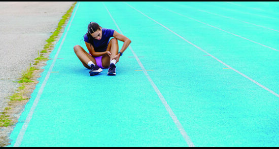 image shows a woman sitting on a running track holding her ankle in pain