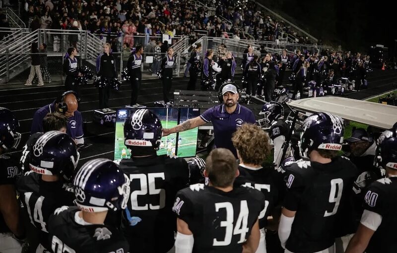 a spanish springs high school football coach points to a video replay screen and talks to a group of high school football players